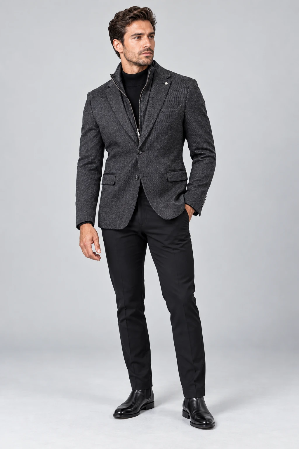 Model wearing a black textured twill sport coat with an integrated zip-up bib, black mock-neck top, black trousers and black shoes on a plain gray studio background.