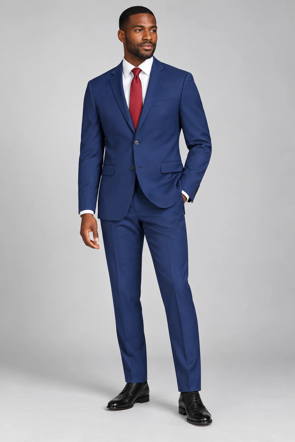 Model wearing a royal blue two-button suit, white shirt, red tie and black shoes on a plain gray studio background.