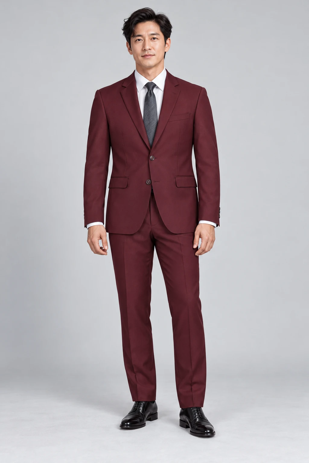 Model wearing a burgundy two-button suit, white shirt, charcoal tie and black leather shoes on a plain gray studio background.